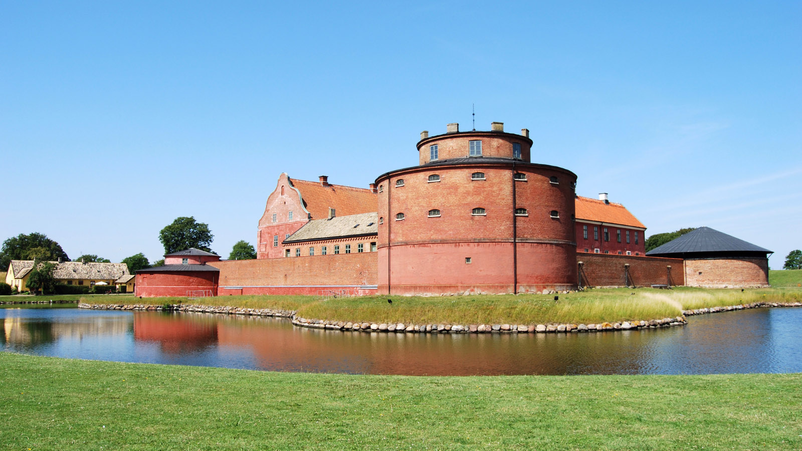 Ett rödbrunt gammalt citadell/ slott. Framför är det vatten och grönt gräs. En blå himmel syns i bakgrunden.