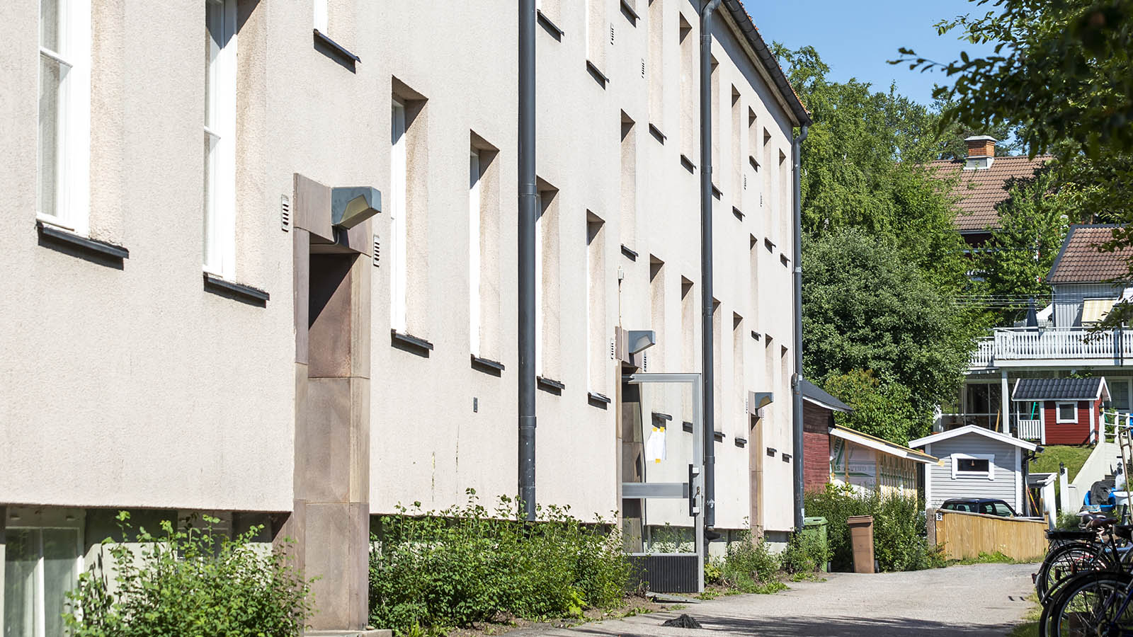 En grå putsad husfasad, ett bostadshus, och i bakgrunden ser man några gamla trävillor på en höjd.