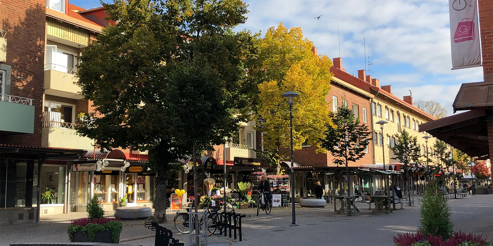 En bild på en gågata i en stad där man ser en blomsterhandel, cyklar som står parkerade, några stora gröna träd.