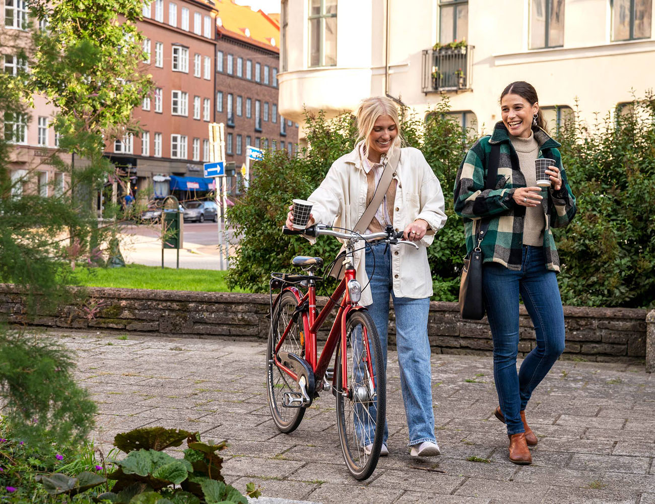 Två tjejer går utomhus med varsin takeaway-kaffekopp i handen och den ena tjejen leder en cykel.