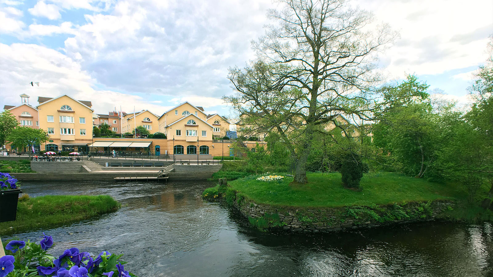 En ström fors där man även ser en grön gräsplätt med träd och gula hus i bakgrunden.