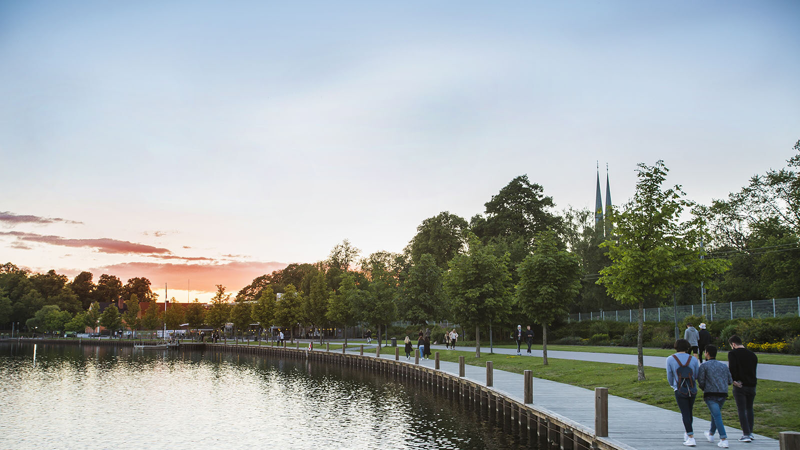 En stor sjö där man ser en gångväg och brygga längs med vattnet som folk går på. En solnedgång nalkas i bakgrunden.
