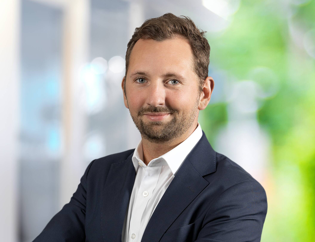 En man med mörkt hår, svart kavaj och vit skjorta står med armarna i kors och ler mot kameran.