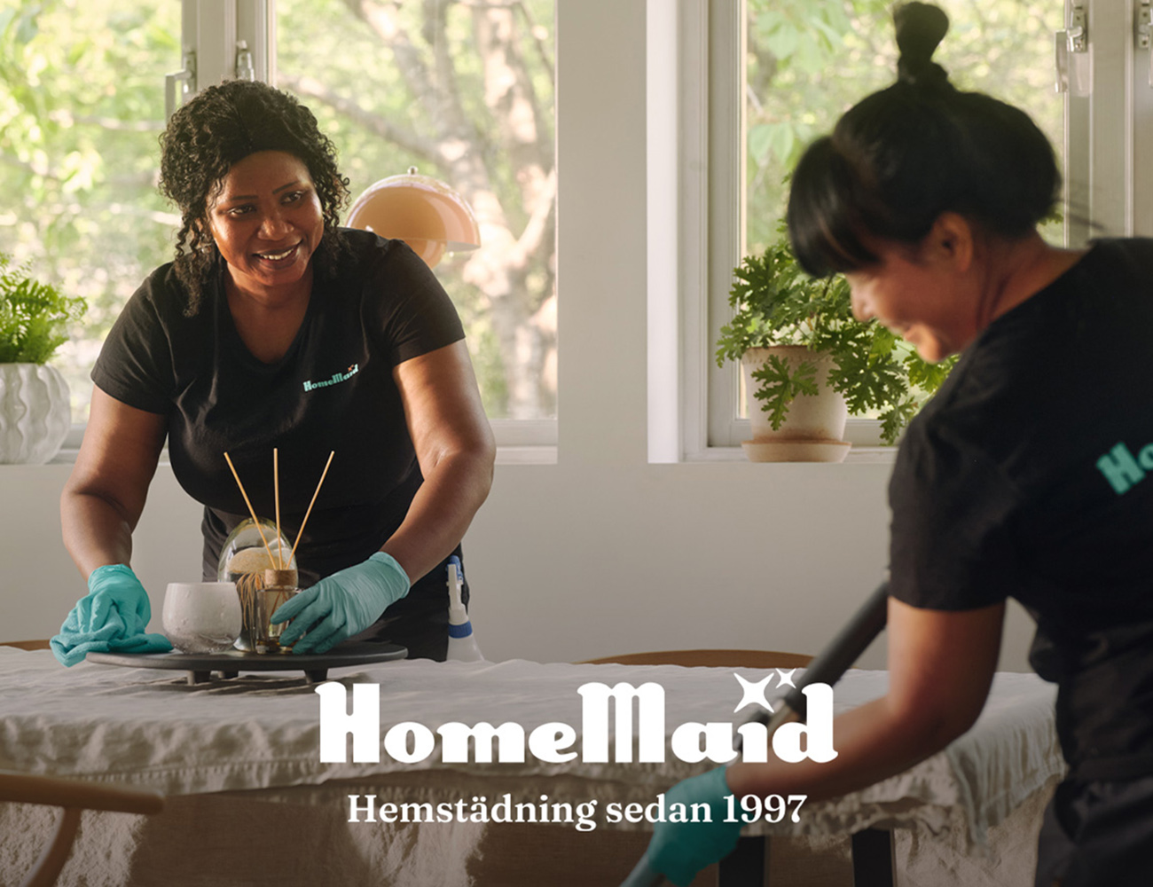 Två kvinnor med blåa handskar på sig, står i ett rum och städar.