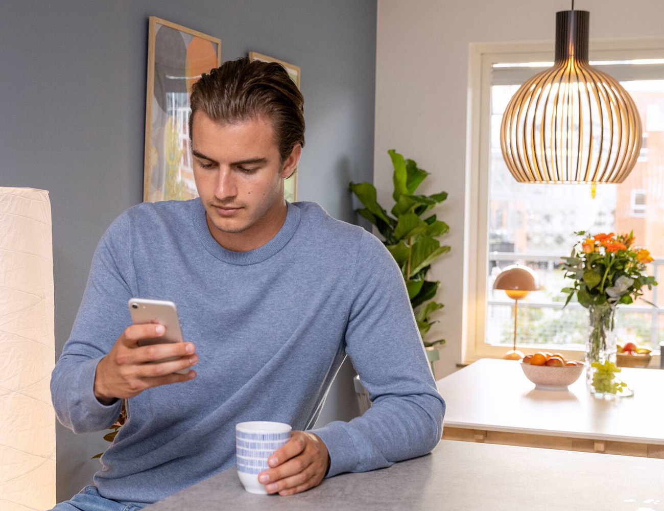 En kille i blå tröja sitter och kollar på sin mobil i handen i ett kök.