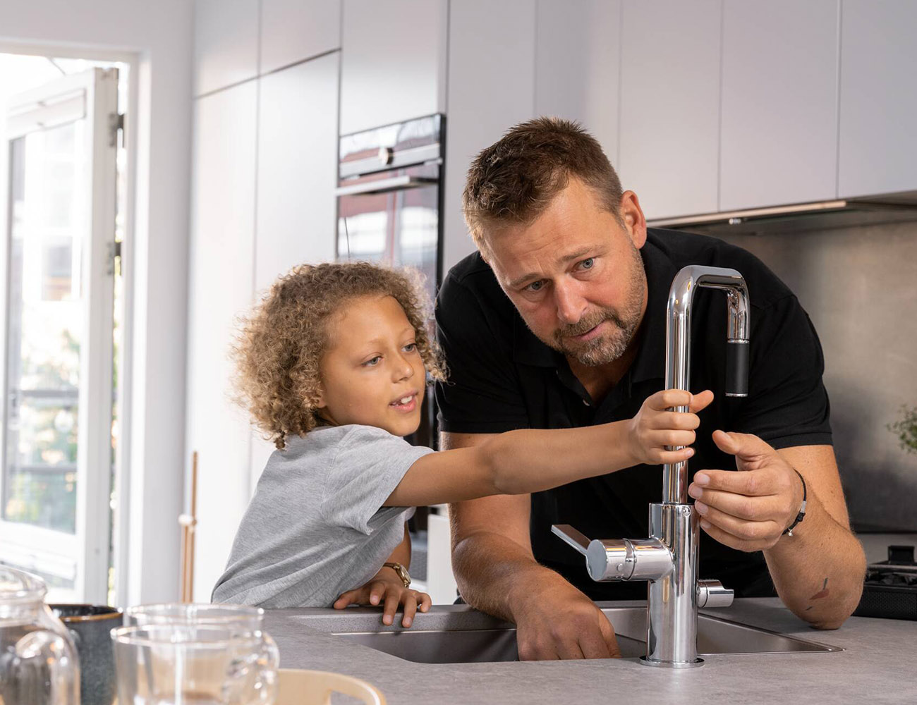 En manlig fastighetsskötare och ett barn står vid en vattenkran i ett kök och försöker få den att funka.