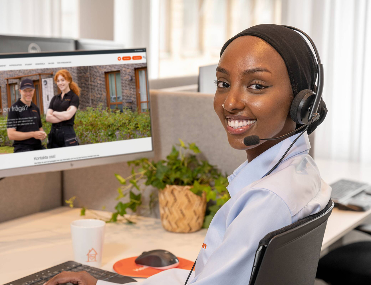 En tjej med blå skjorta och headset sitter framför en dator i en kundtjänst och ser glad ut.