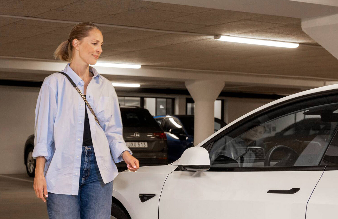 En kvinna med blå skjorta går fram till en vit parkerad bil i Heimstadens garage.
