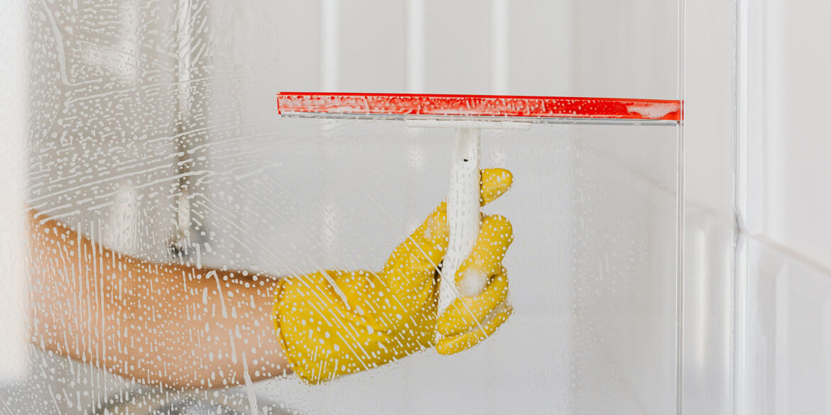 A white and orange shower scraper used on a glass wall. A hand with a yellow glove on pulls the scraper on the shower wall.