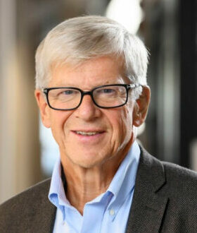 A man with grey hair and black glasses, a blue shirt and a dark brown jacket smiles at the camera