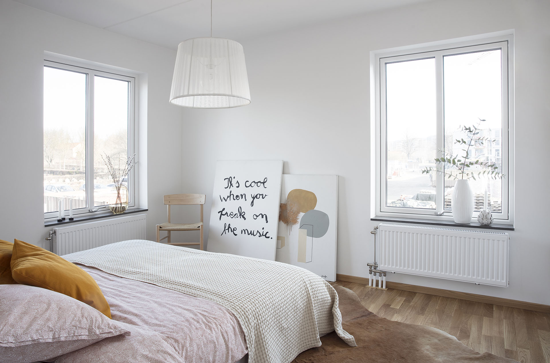 A large white bedroom with a big bed in white and pink and two windows. There is a white lamp in the ceiling.