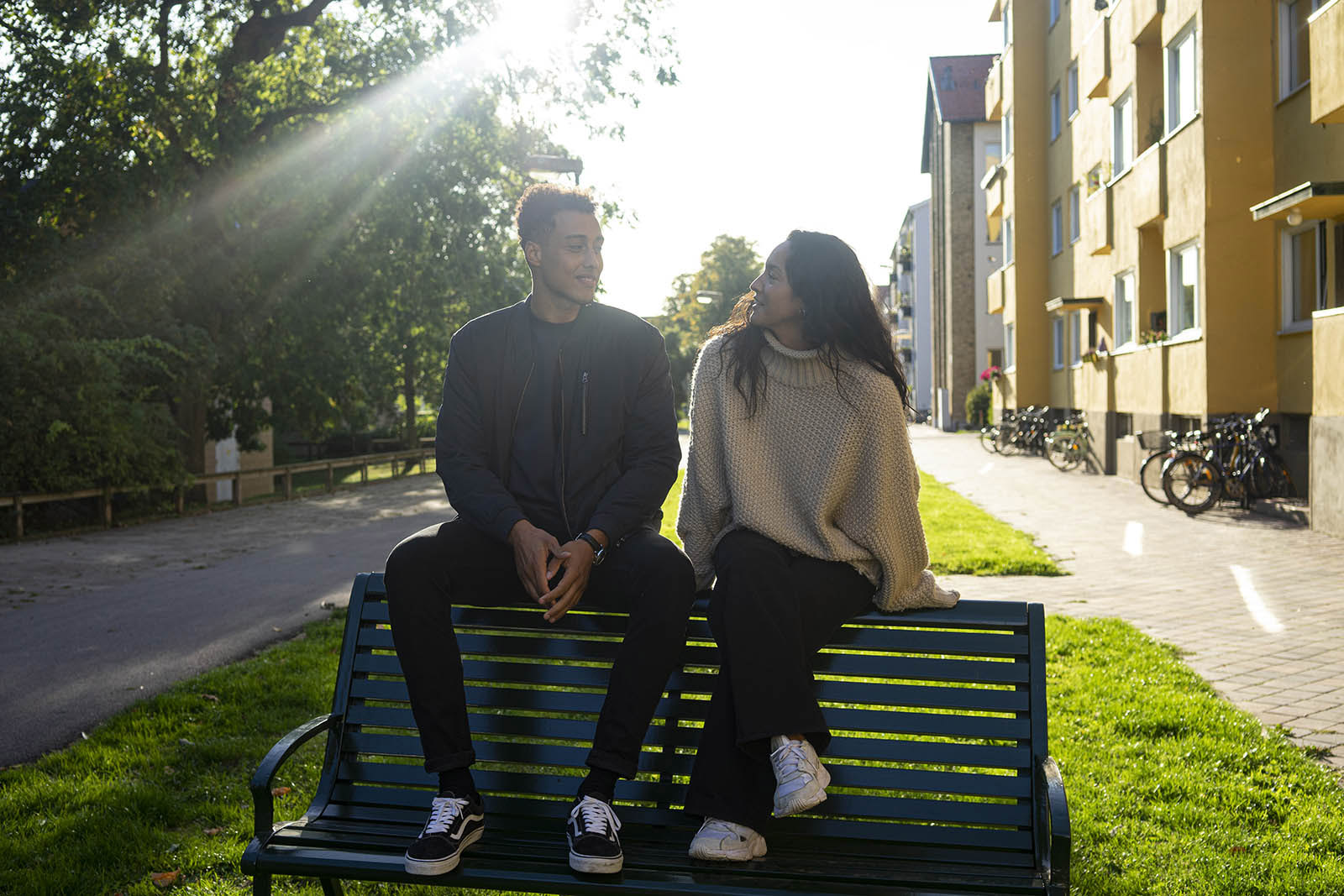 Two people are sitting on the backrest of a park bench in front of a block of flats.