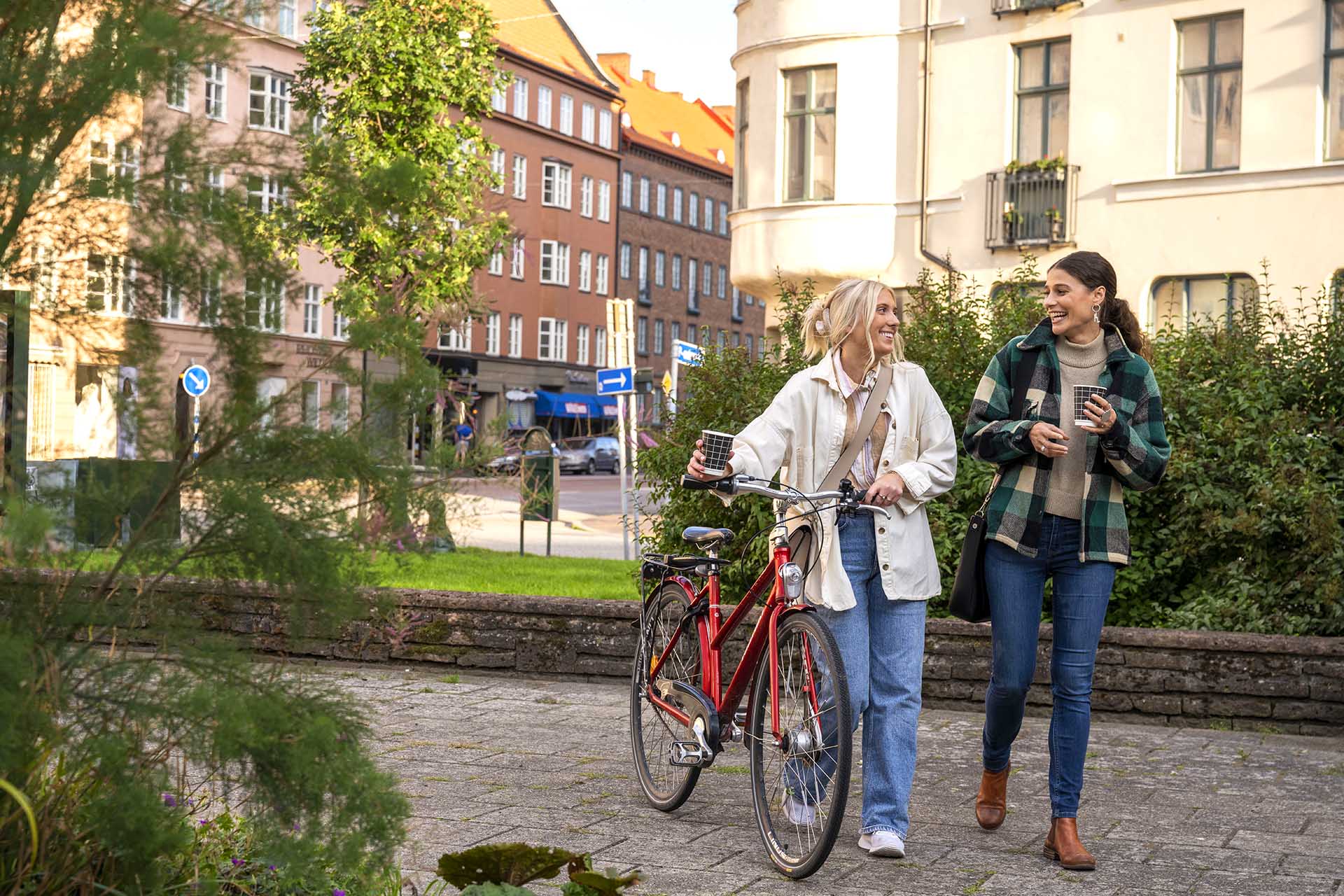 Two women are walking through a park. One of them is holding onto her red bike. They are talking and laughing,