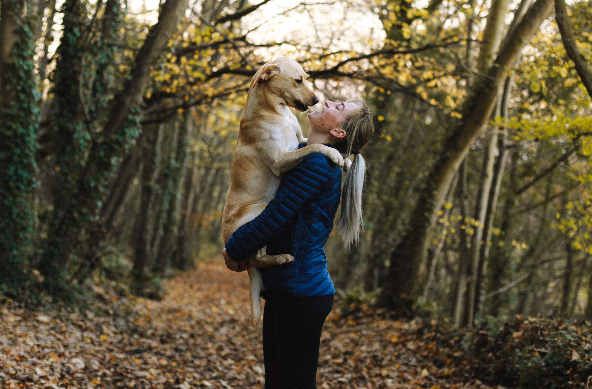 A woman with long hair is standing in the woods with a big dog in her arms. They are looking happily at each other.