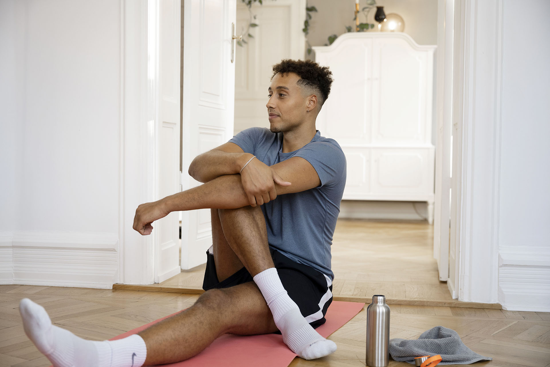 A man with short dark hair and a grey shirt and black shorts is sitting on the floor on a yoga mat. Next to the carpet is a bottle of water and a towel.