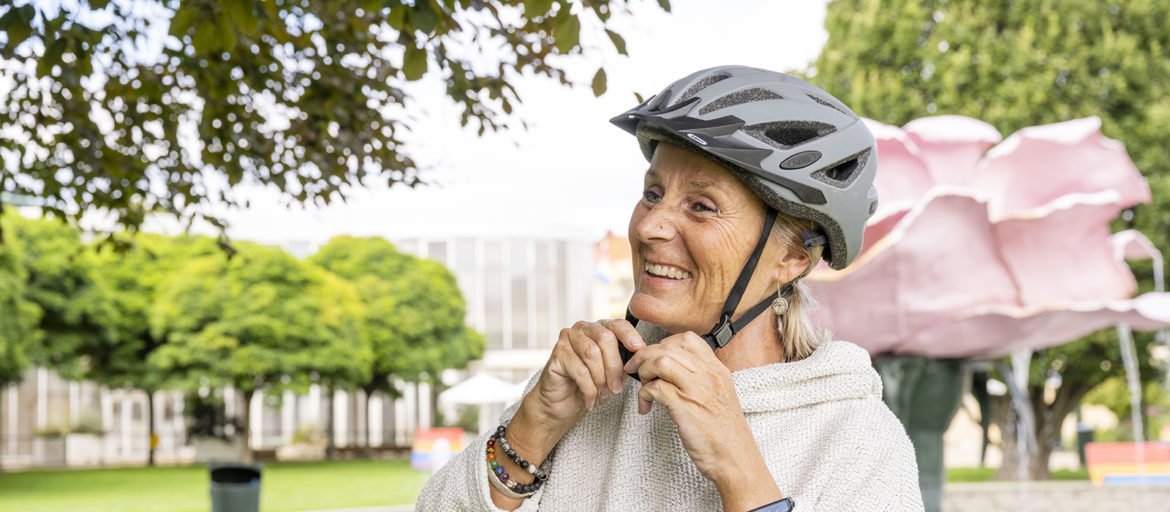 A woman in a grey jumper is putting on a black bicycle helmet. There are green trees in the background.