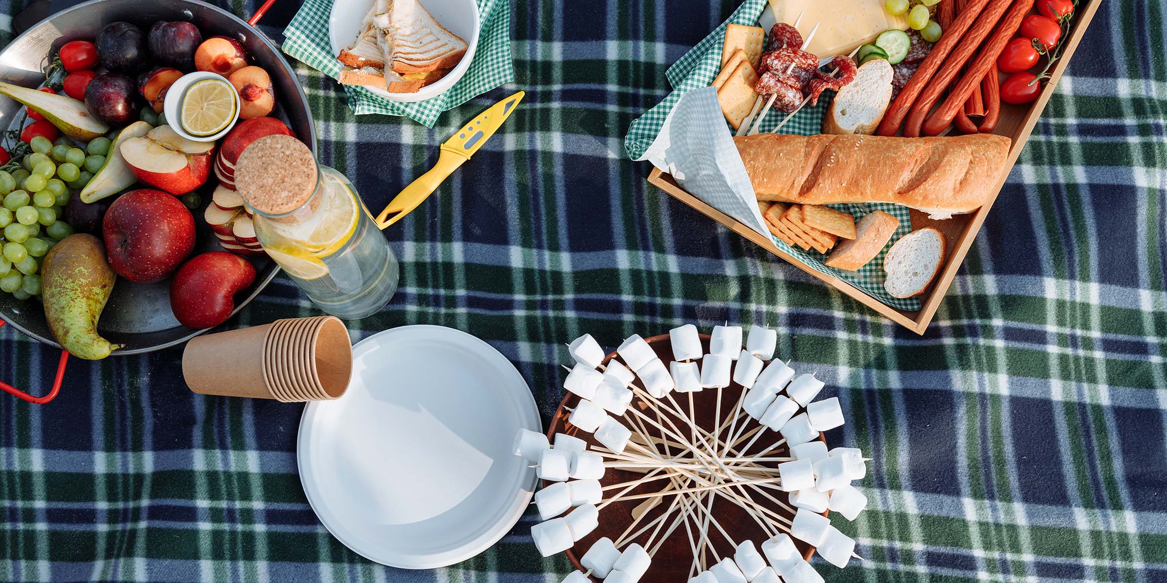 A checkered blanket with a plate of marshmallows, a basket of bread and fruit and some plates.
