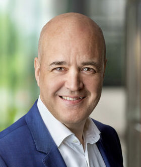 A bald man in a white shirt and blue jacket smiles at the camera