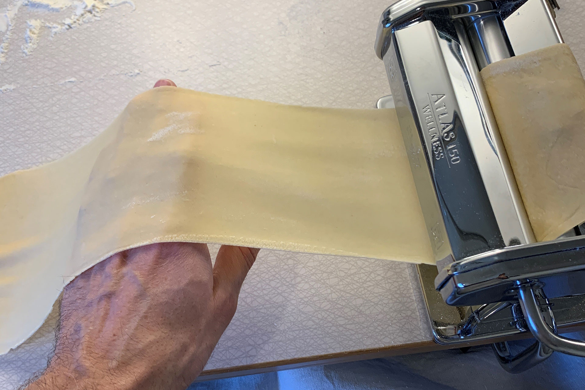 A hand is holding pasta dough to run it through a pasta machine.