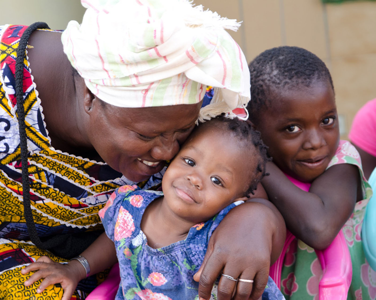 A person in a white turban is hugging a small child in a blue dress. Antoher child is hanging on to both of them.
