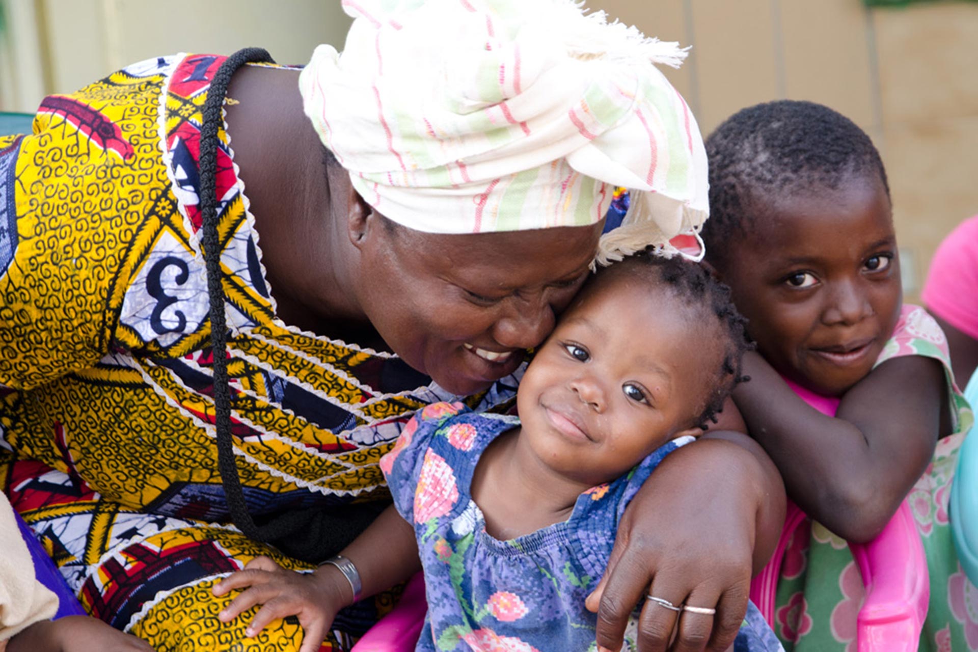 A woman in a colourful dress and white headdress is hugging two children