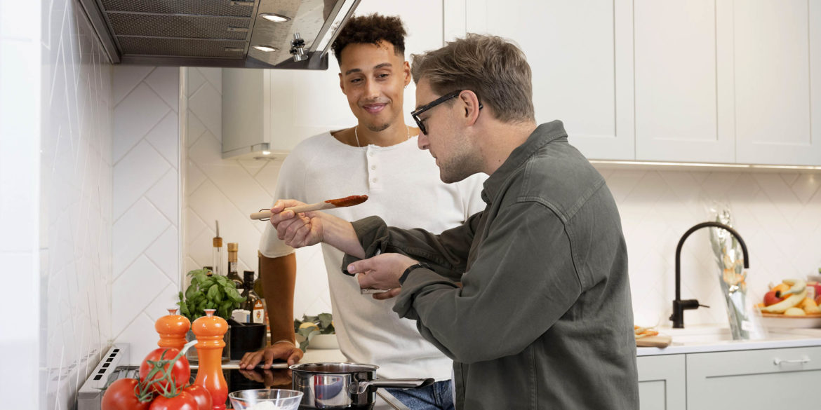 Två killar står i ett kök och den ena smakar av något som han lagar på spisen