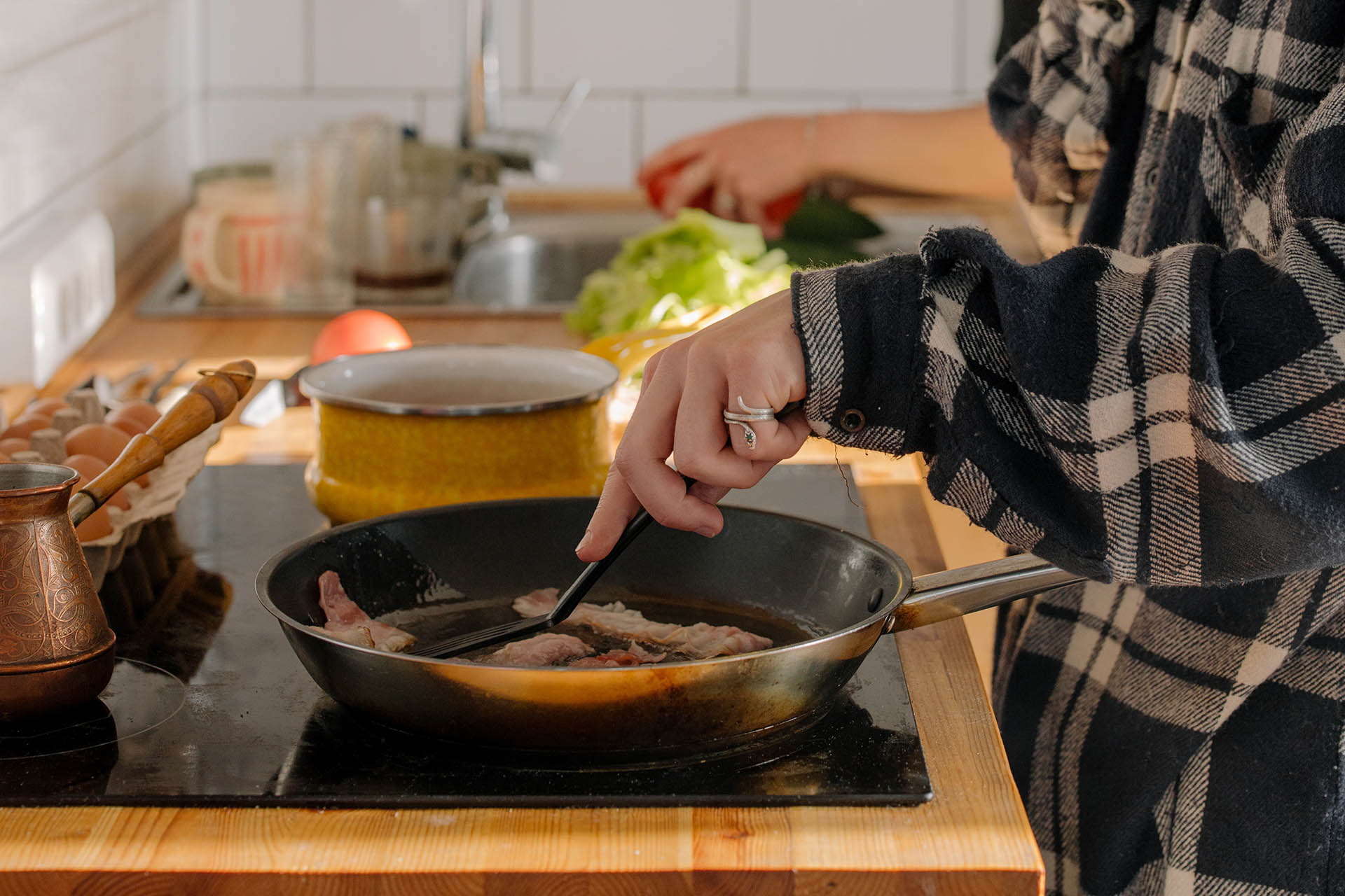 A person is standing by the stove in a checkered shirt, frying bacon in a frying pan.