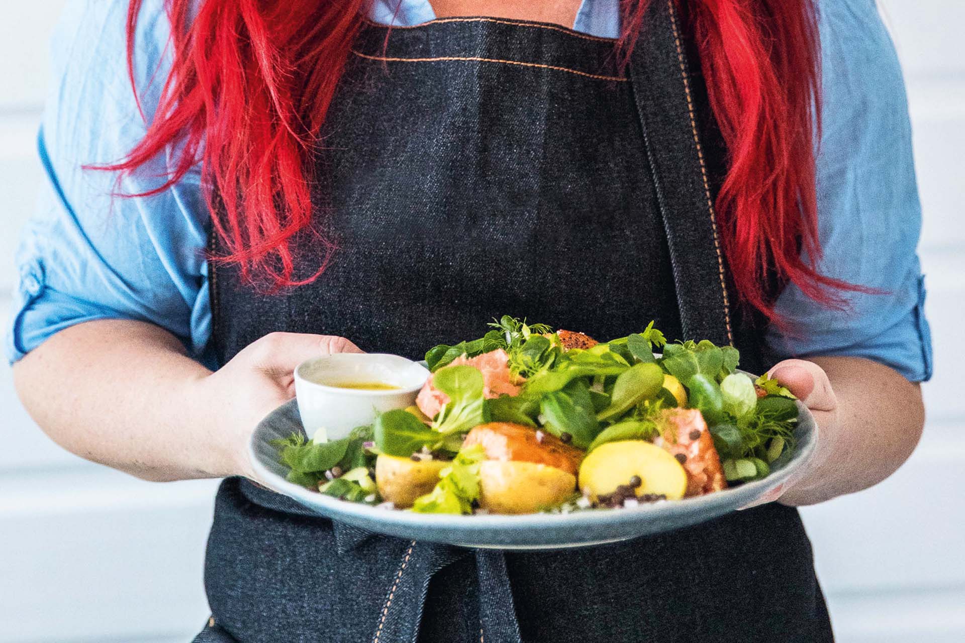 A woman with red hair and an apron is holding a plate of salmon, potatoes and green salad in her hands.