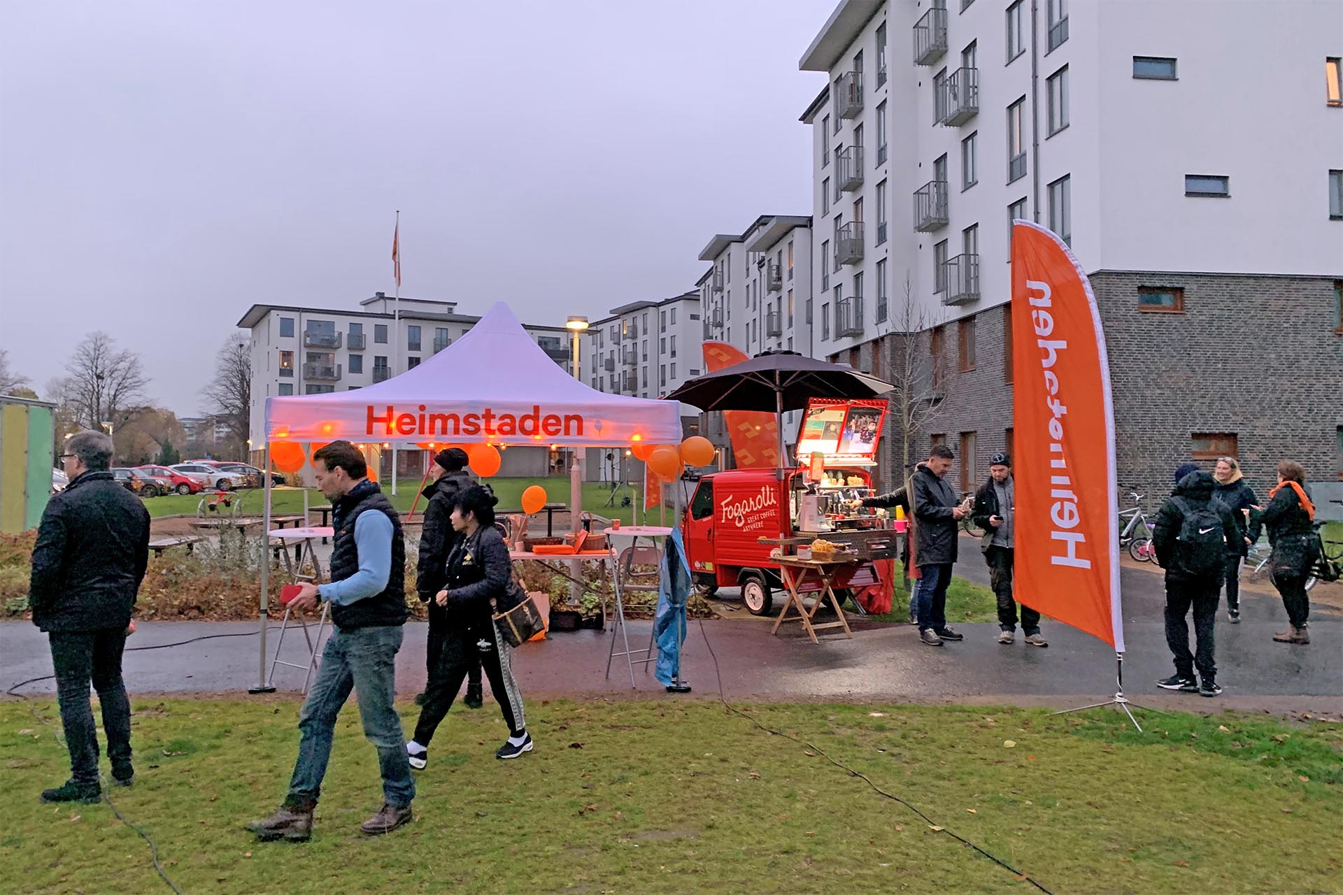 A tent and a flag with Heimstaden logotypes in front of a grey block of flats. People are moving around.