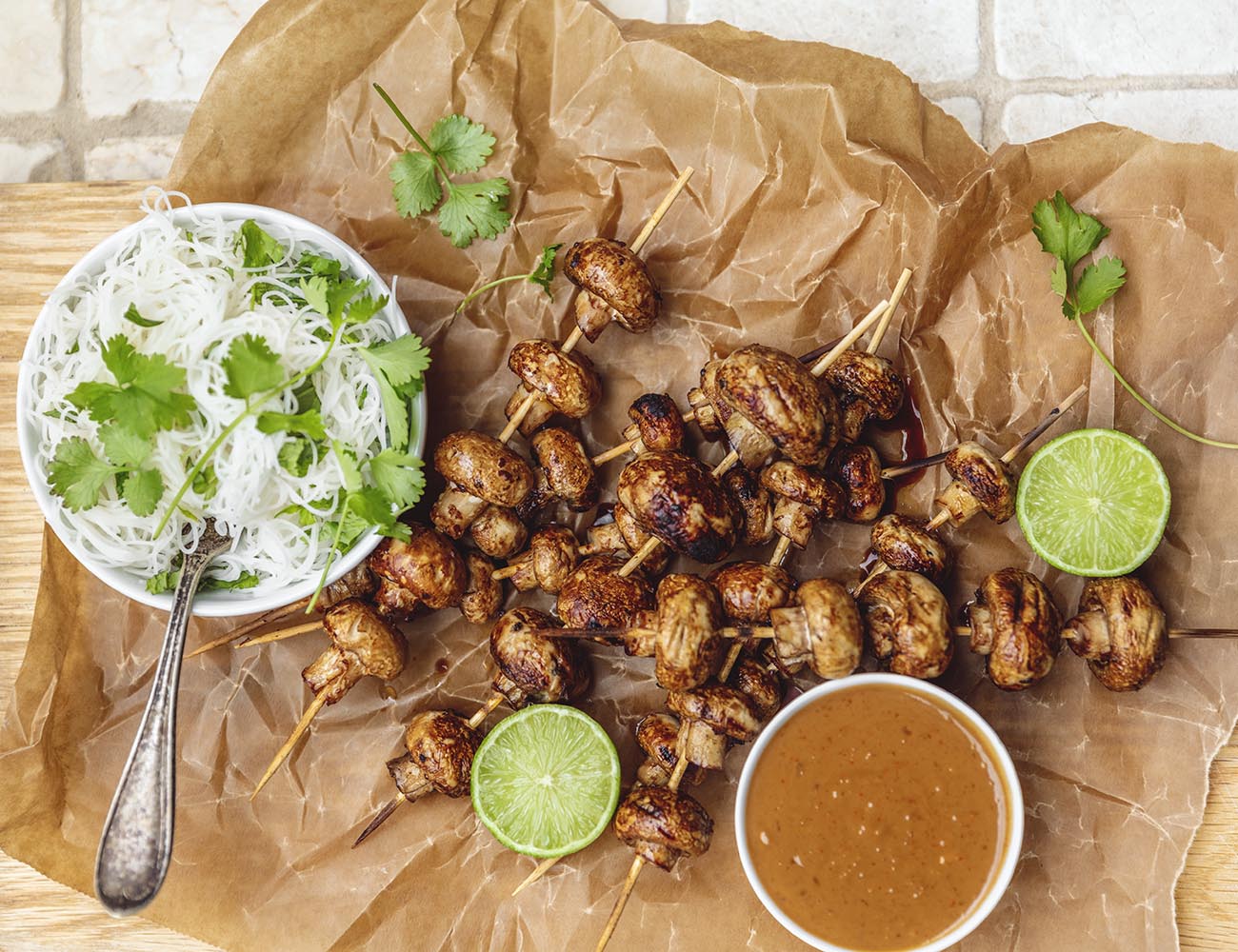 Mushroom skewers are lying on a piece of paper with lime, a bowl of noodles and a bowl of peanut sauce.