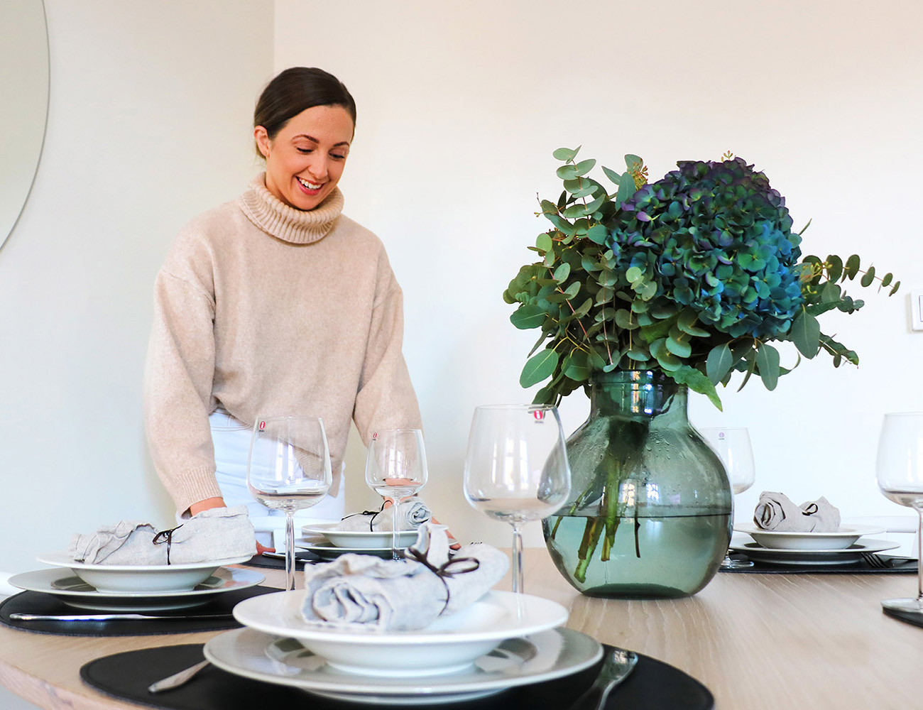 A girl with dark hair and a beige thick polo shirt stands at a dining table and tablecloths. On the wooden table you see a large green glass vase with cut flowers and porcelain in white and black.