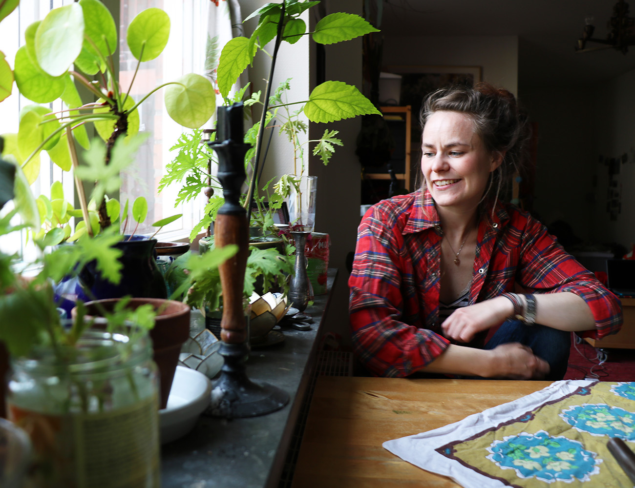 A woman with dark hair and a red checkered shirt sits at her kitchen table and looks at her green plants in the window frame.