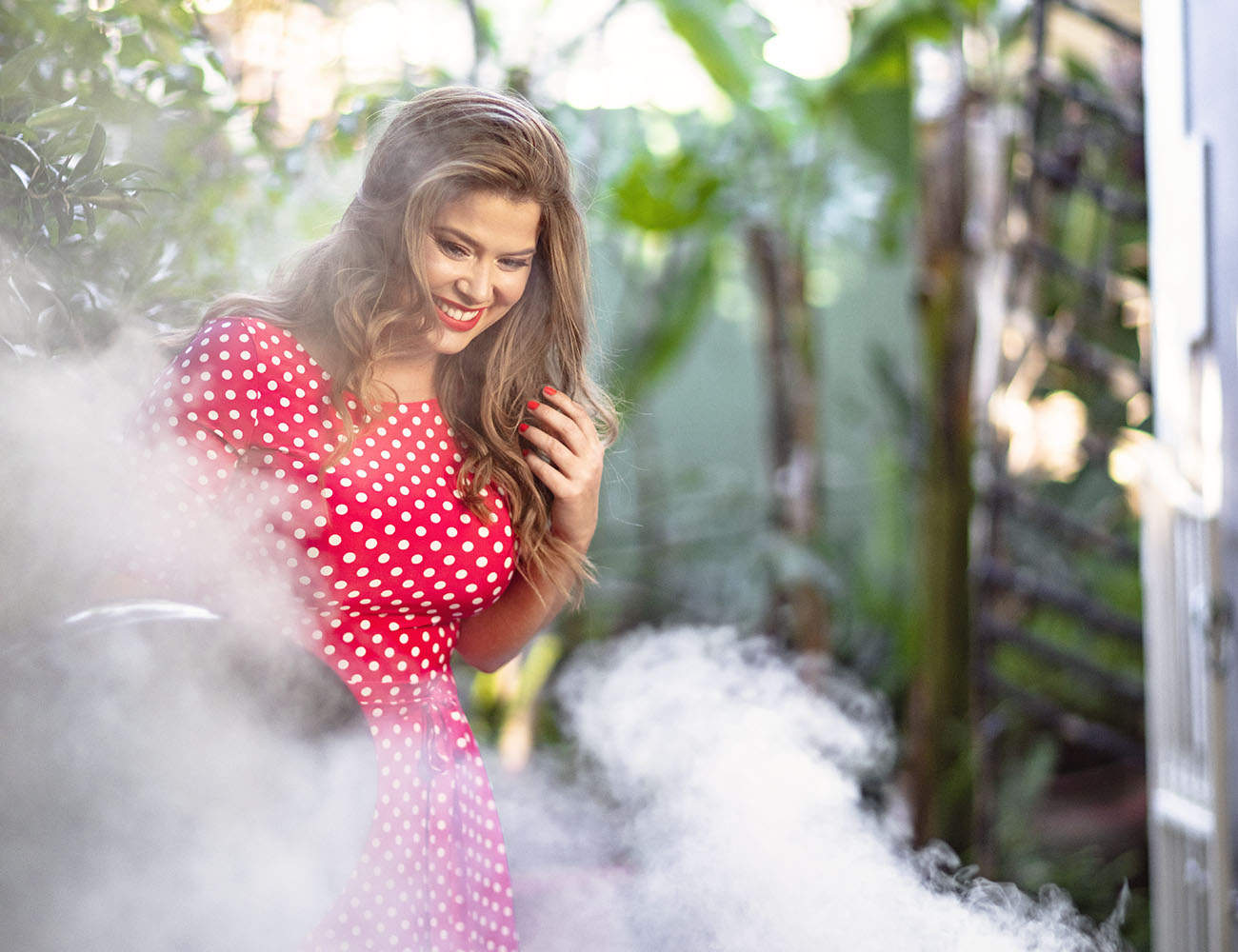 A woman in a dotted pink dress is standing next to a grill with lots of smoke coming out of it.