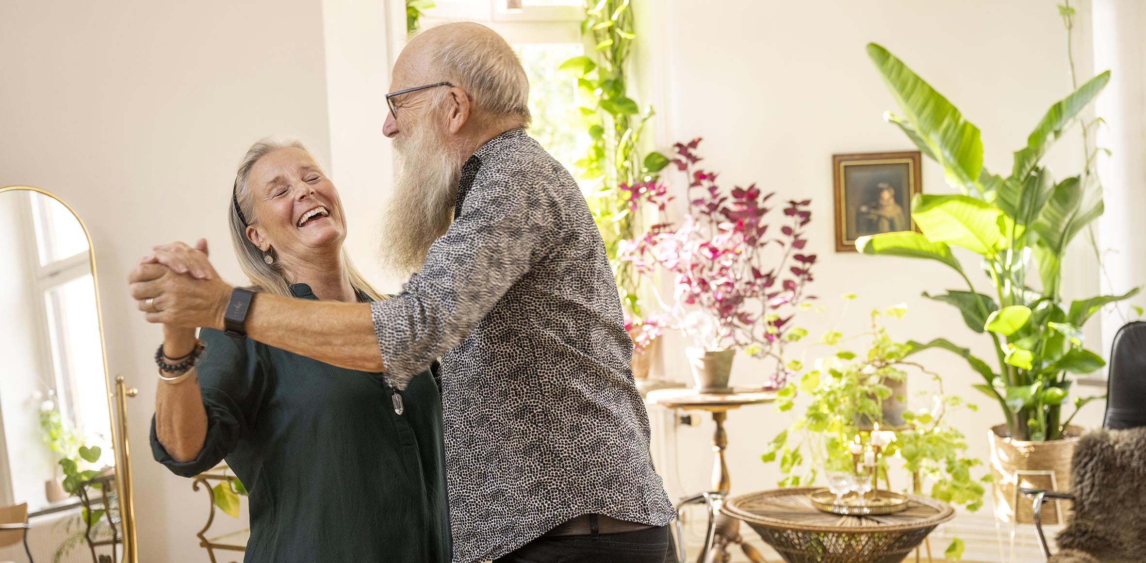An older couple are dancing in their living room with green plants and white walls