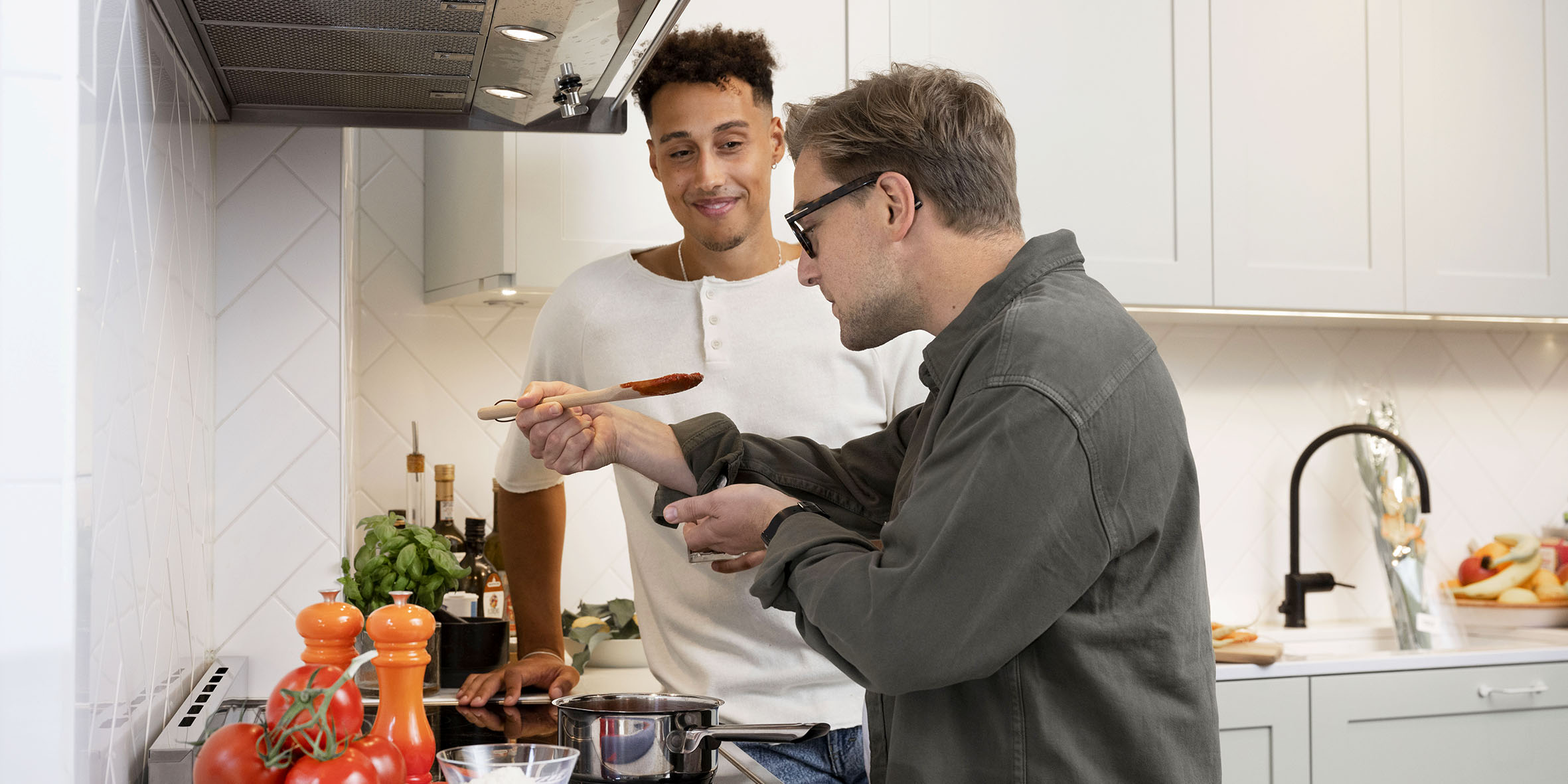 Two men are cooking in a kitchen, standing next to the stove and trying a sauce that is boiling in a pot.