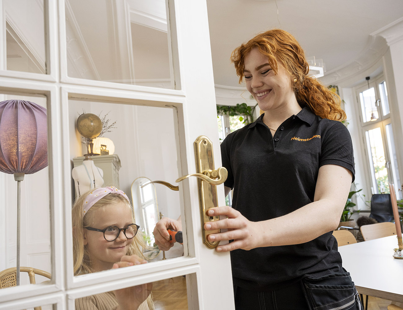 A red-haired woman in a Heimstaden uniform is screwing on a door handle while a little girl watches.