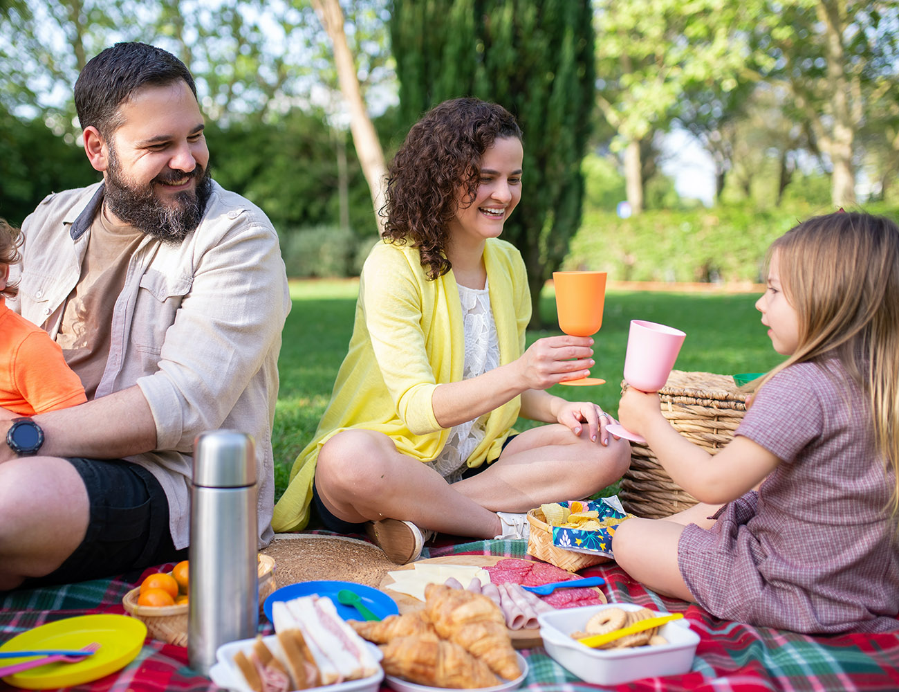 A man, a woman and a child are sitting on a picnic blanket out on a lawn. On the blanket it is served with a thermos, various bowls with food and bread in it.