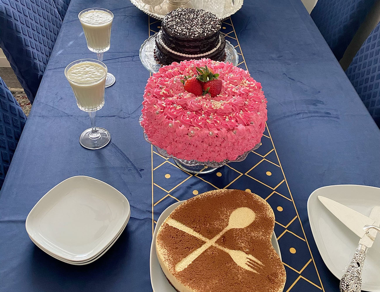 A table with a blue tablecloth where there are plates, glasses with some light frothy drink in and four dishes with different kinds of cakes.