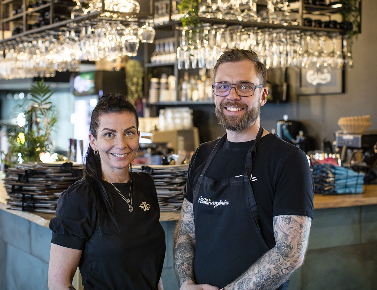 A woman and a man with black aprons are standing in front of a bar with lots of glasses hanging from a shelf.