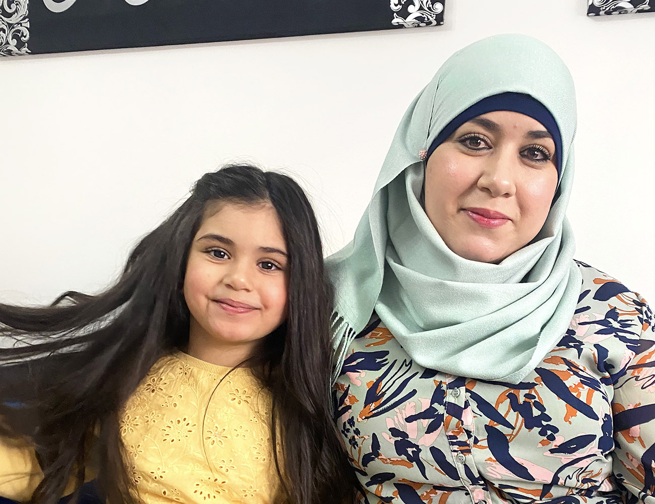 A woman with a light blue headdress and a patterned dress in blue and orange is sitting on a sofa with a little girl wearing a yellow sweater.