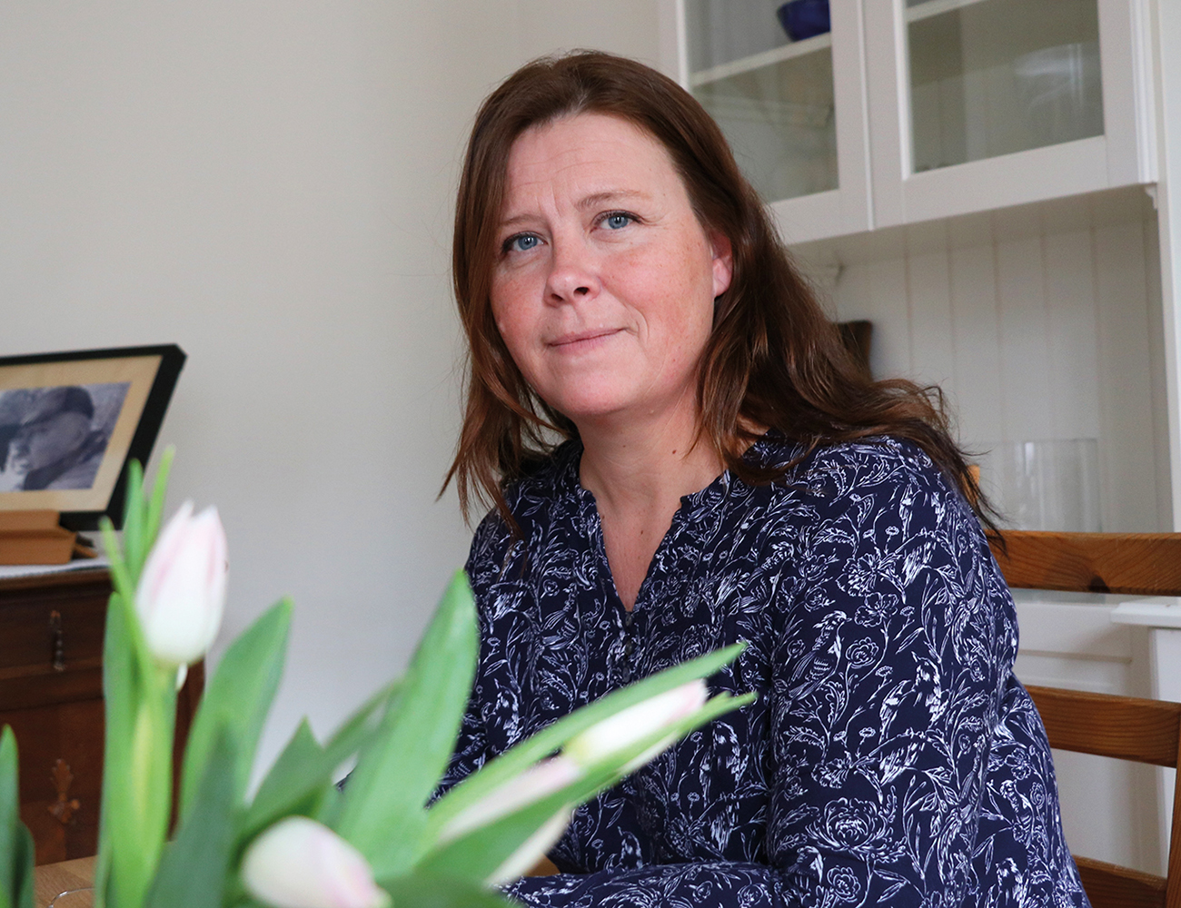 A woman in dark brown hair is sitting on a kitchen chair with a bouquet of tulips in front of her.