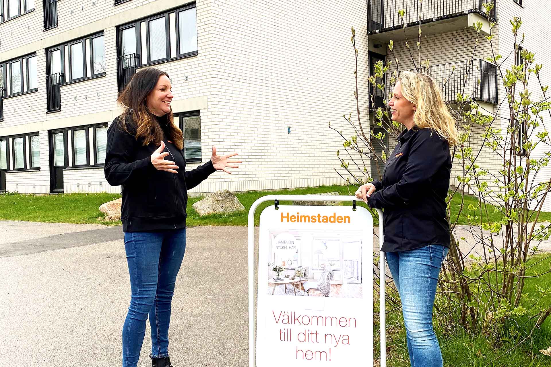 Two women stand in front of a bright residential building and talk to each other. One of them leans against a sidewalk speaker that says Welcome to your new home.