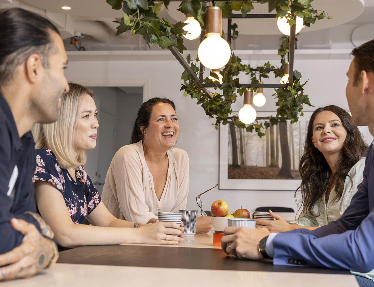 Three women and two men sit at a table and drink coffee while talking and smiling at each other. A brass lamp hangs from the ceiling.