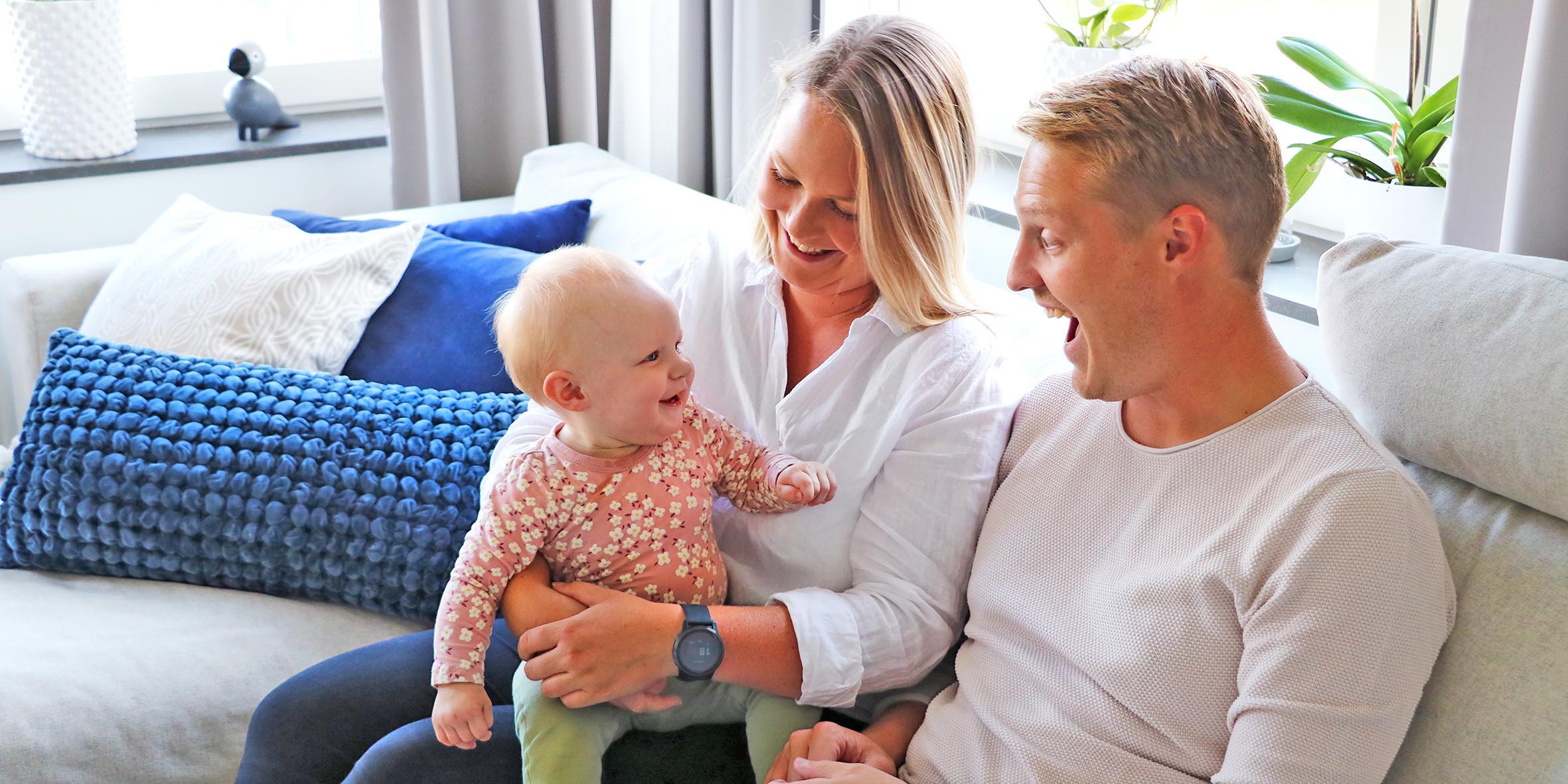 A young couple is sitting on a white sofa and the woman has a child in her arms. The man makes a face and the child laughs.