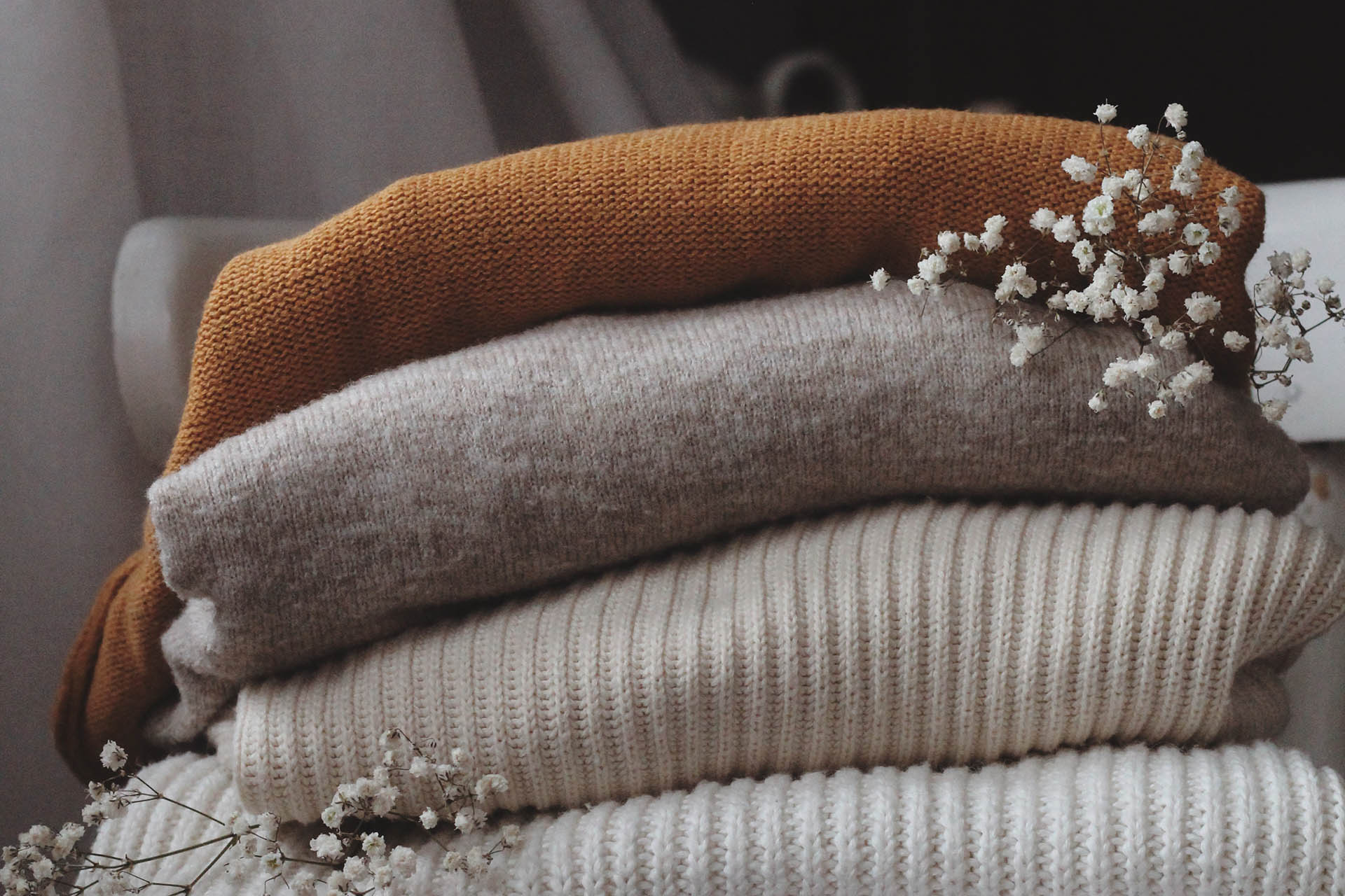 Four thick sweaters in white, beige and rust brown that are folded nicely. Two white flower twigs lie on the clothes for decoration