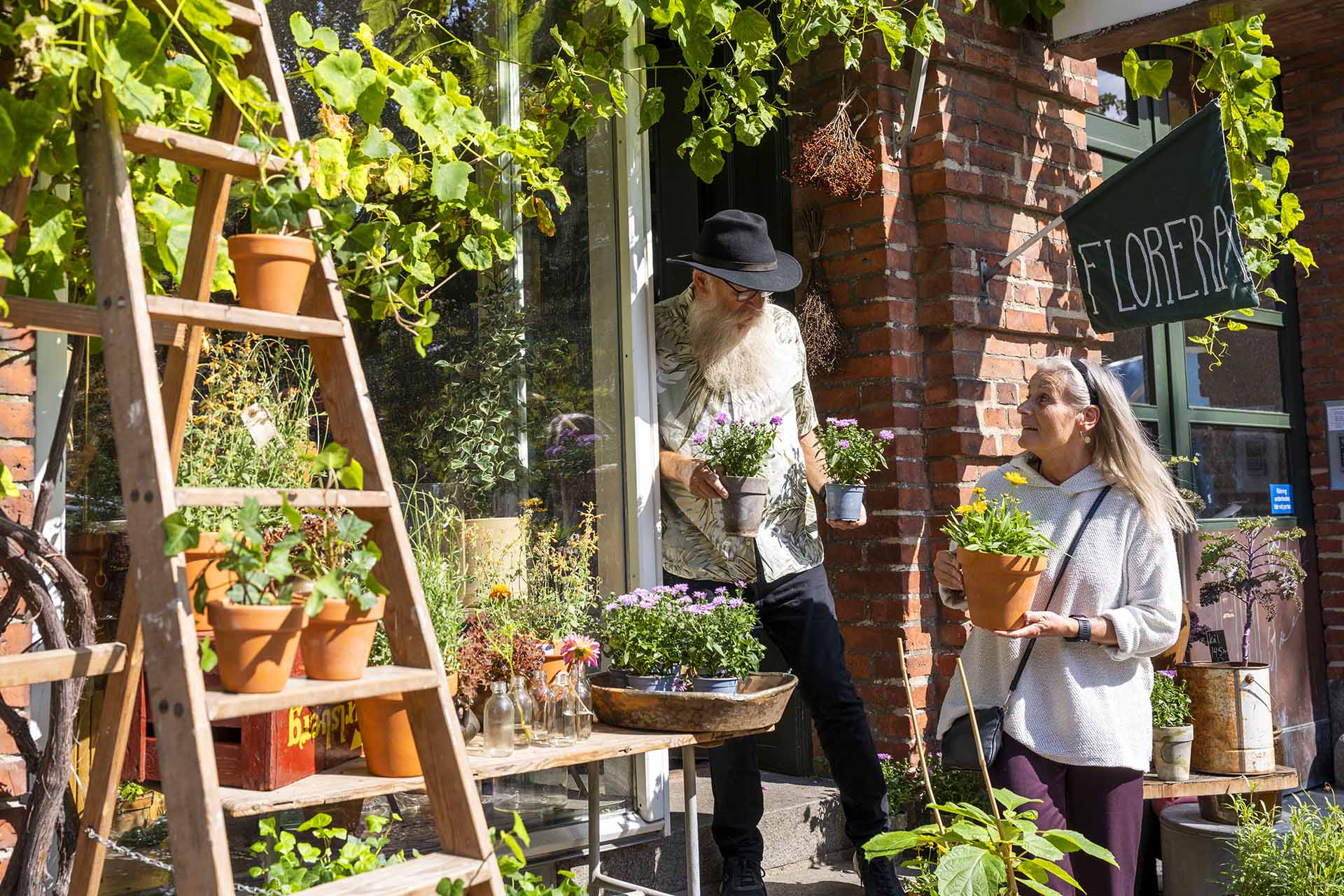 A man with a white beard and black hat stands in the doorway of a flower shop with a woman. They hold potted plants in their hands.