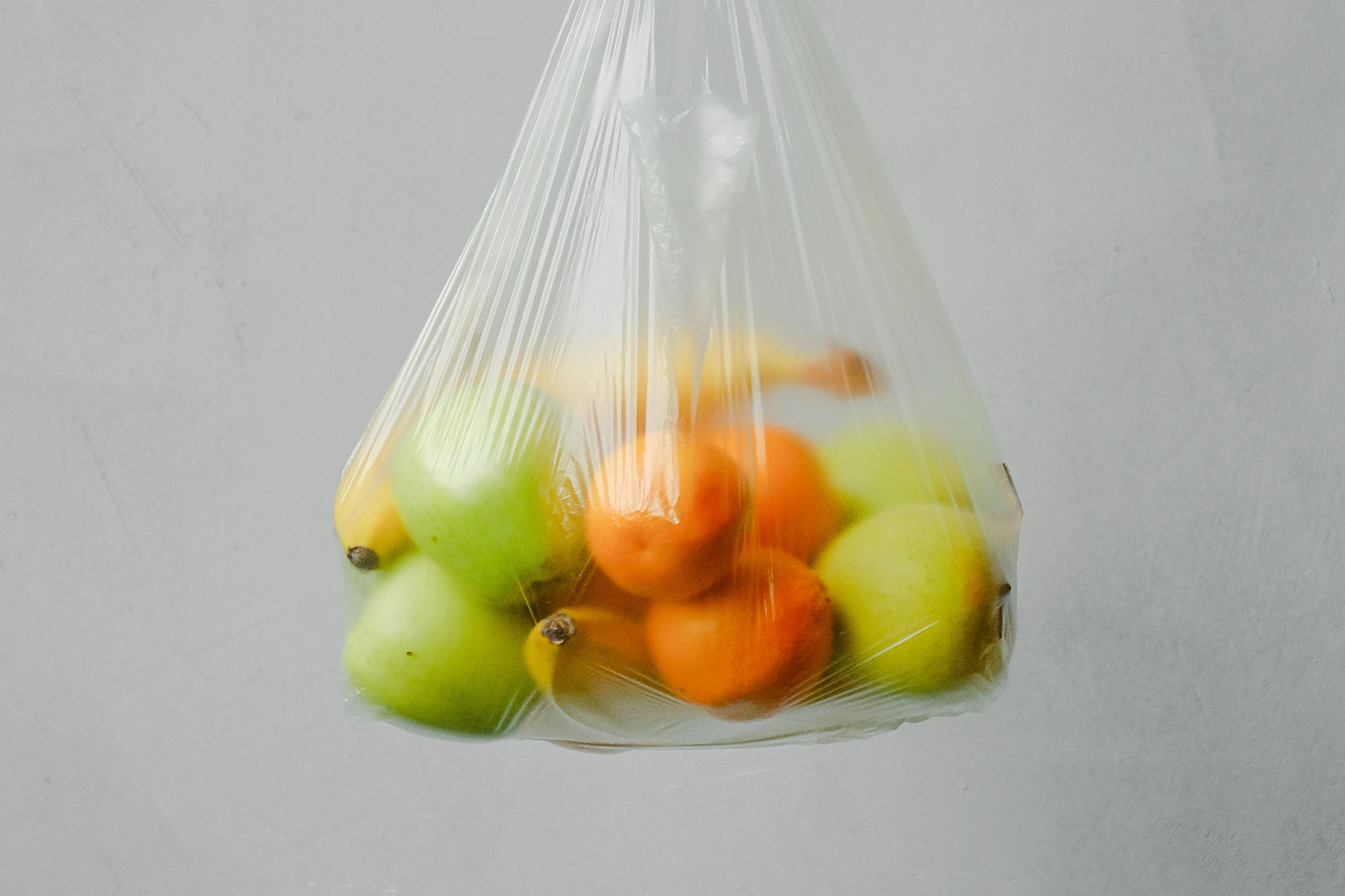 A transparent plastic bag that hangs in the air. It contains tangerines, green apples and yellow bananas.