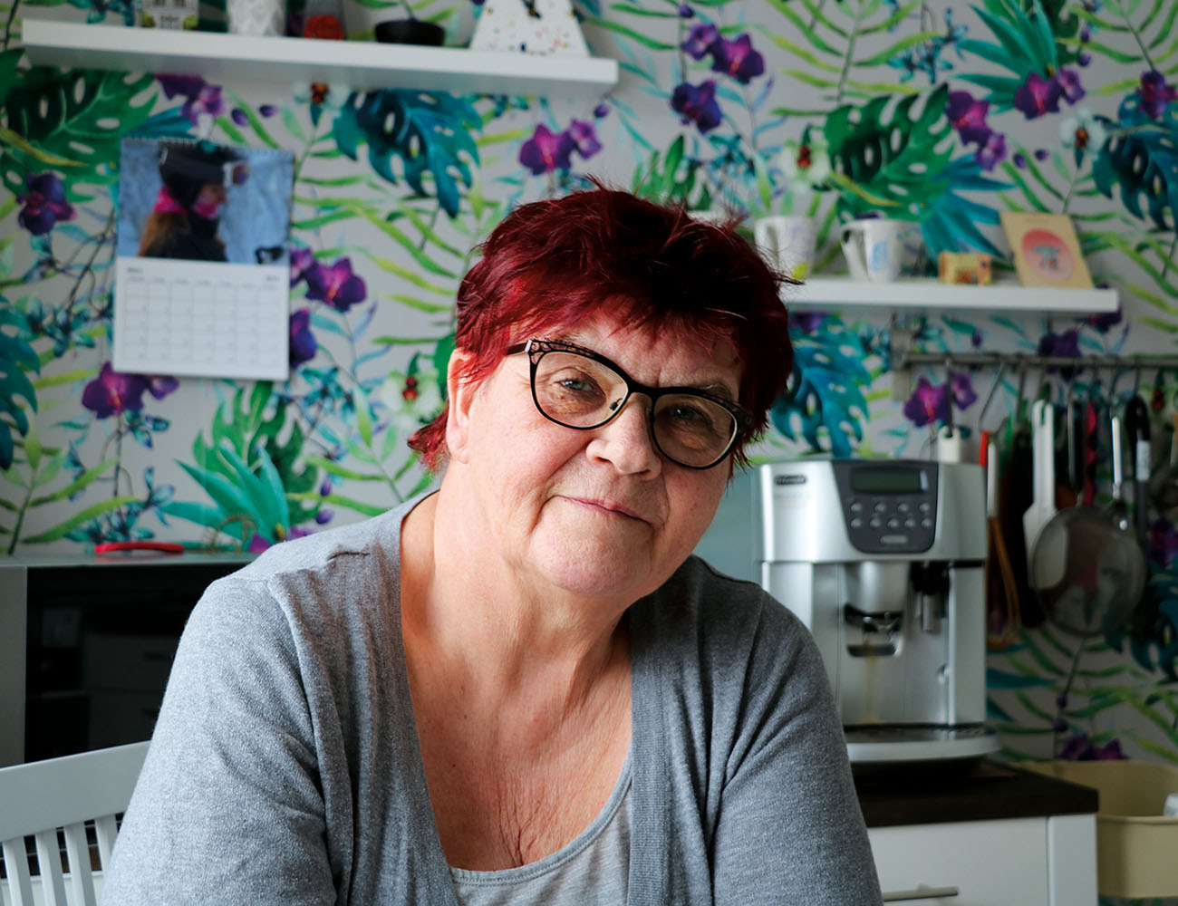 A woman with short red hair and glasses is sitting in a kitchen where you see a colorful and patterned wallpaper behind her.