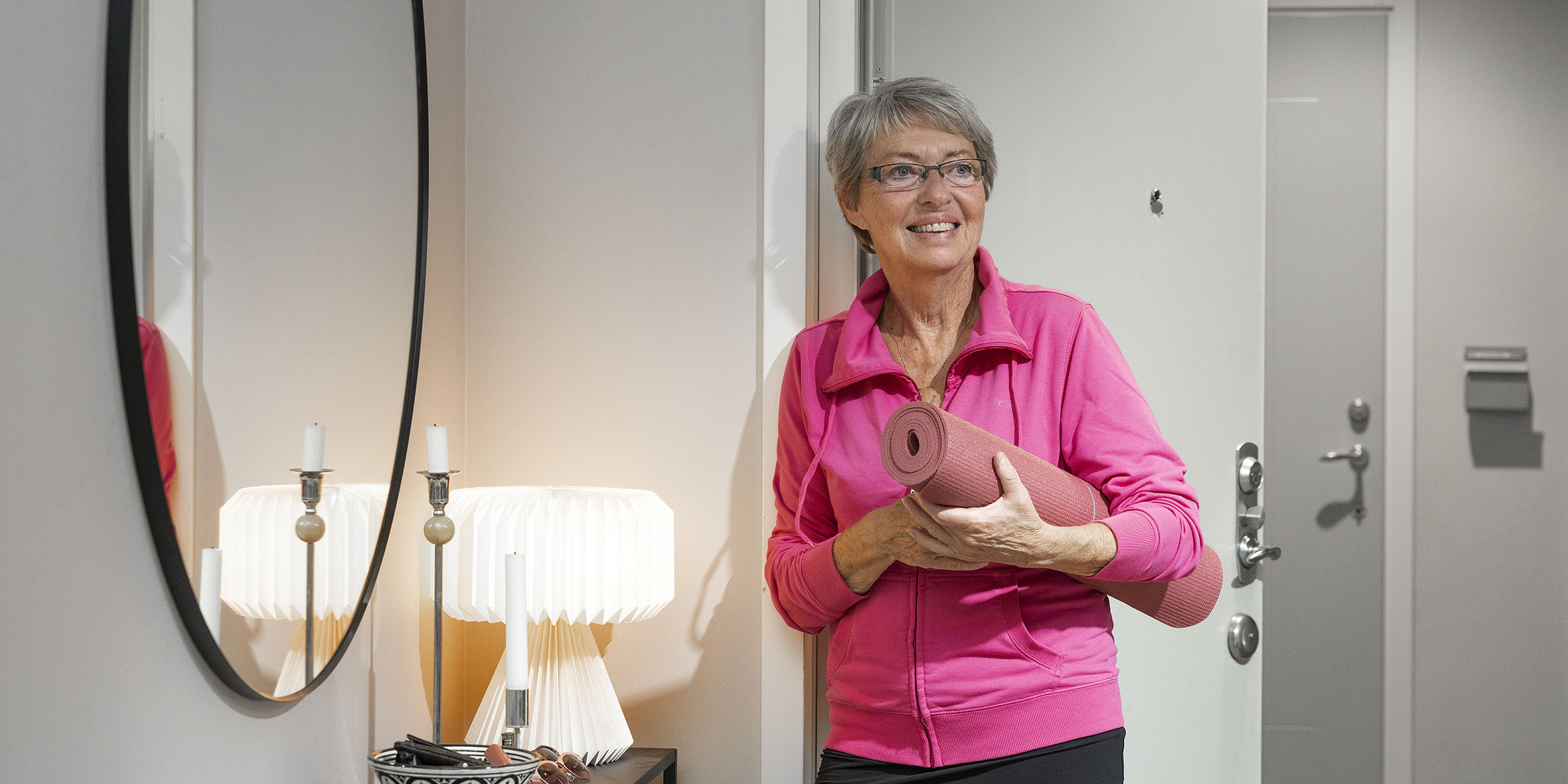 En äldre kvinna med en rosa träningsjacka står med en ihoprullad roa yogamatta under armen. Hon står och lutar sig mot en dörrpost och man ser en mörk byrå, en vit bordslampa som är tänd och en del av en spegel på väggen.