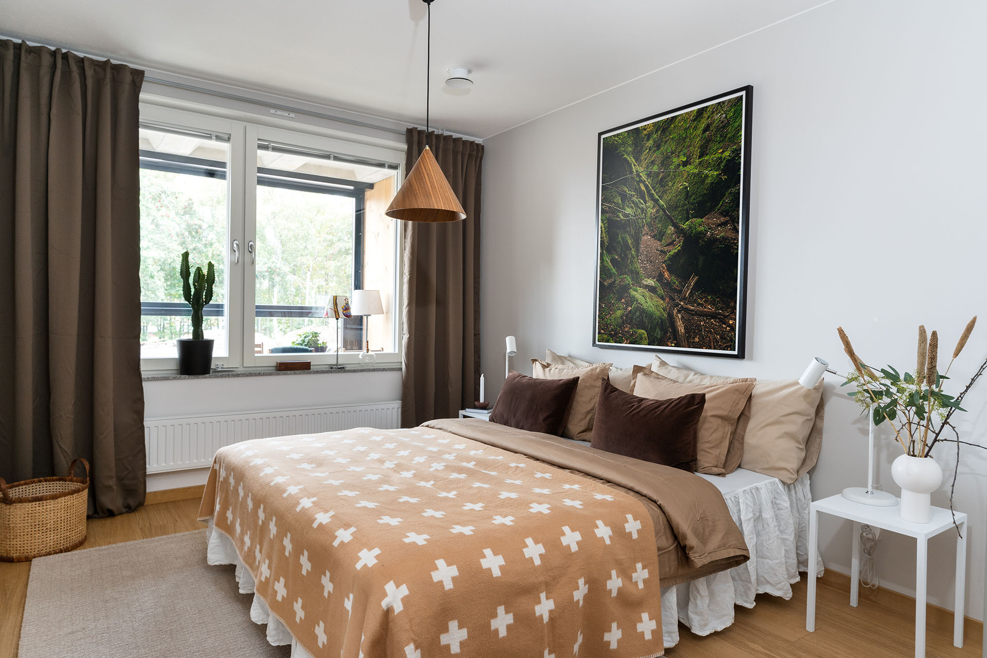 Ett sovrum med en säng i mitten som har en brunt överkast med ett korsmönster. Brungröna långa gardiner i fönstret och en tavla med skogsmotiv på väggen.